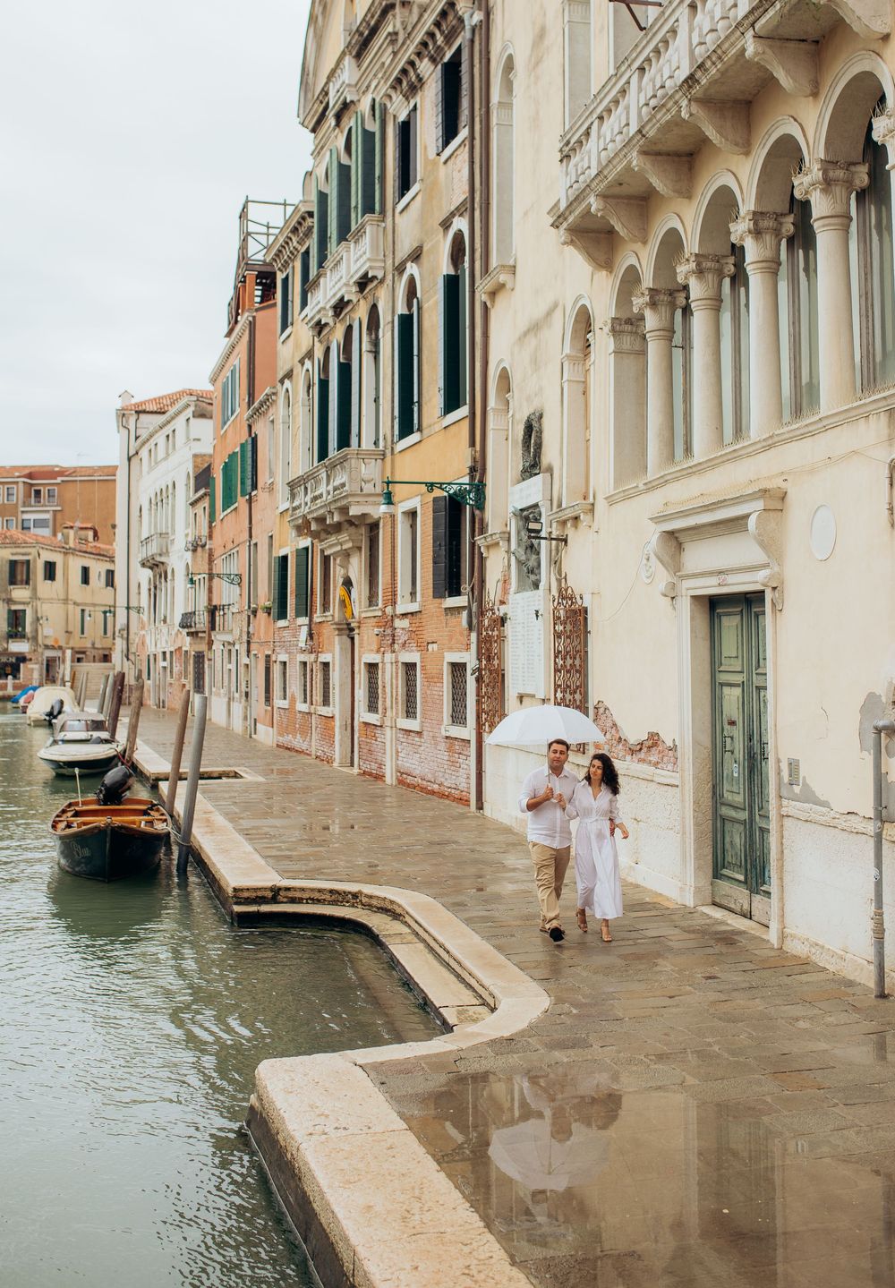 A scenic Venice canal with historic architecture and a couple under a white umbrella walking along the waterfront path.
