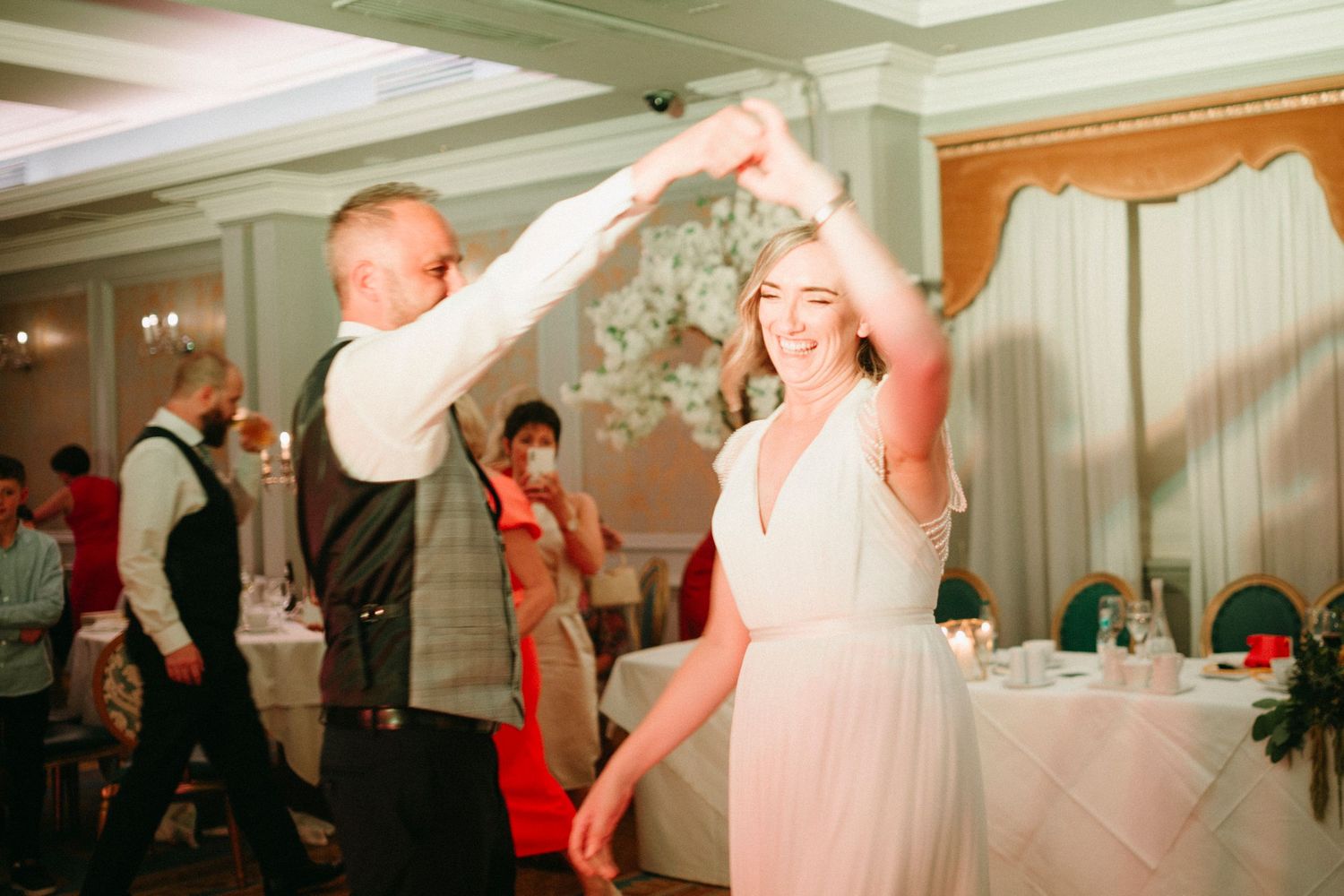 A couple dances together in a white reception hall during a wedding celebration, captured in black and white photography.