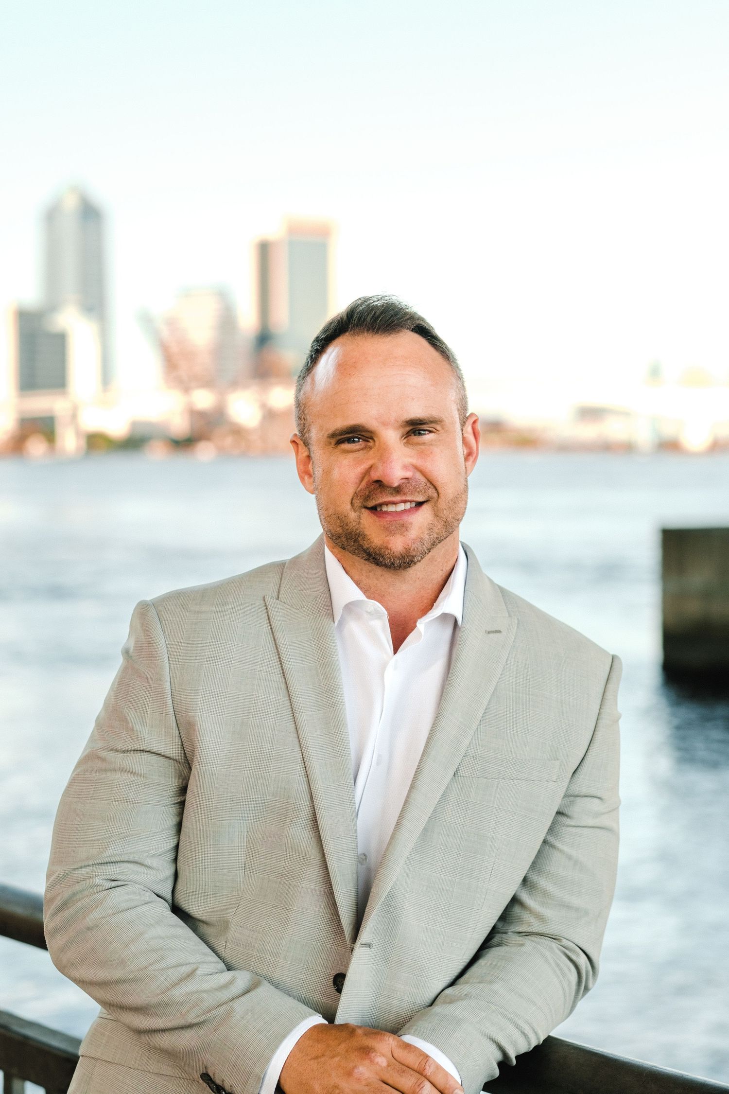Professional headshot of a smiling man in a beige suit by a waterfront with a city skyline backdrop.