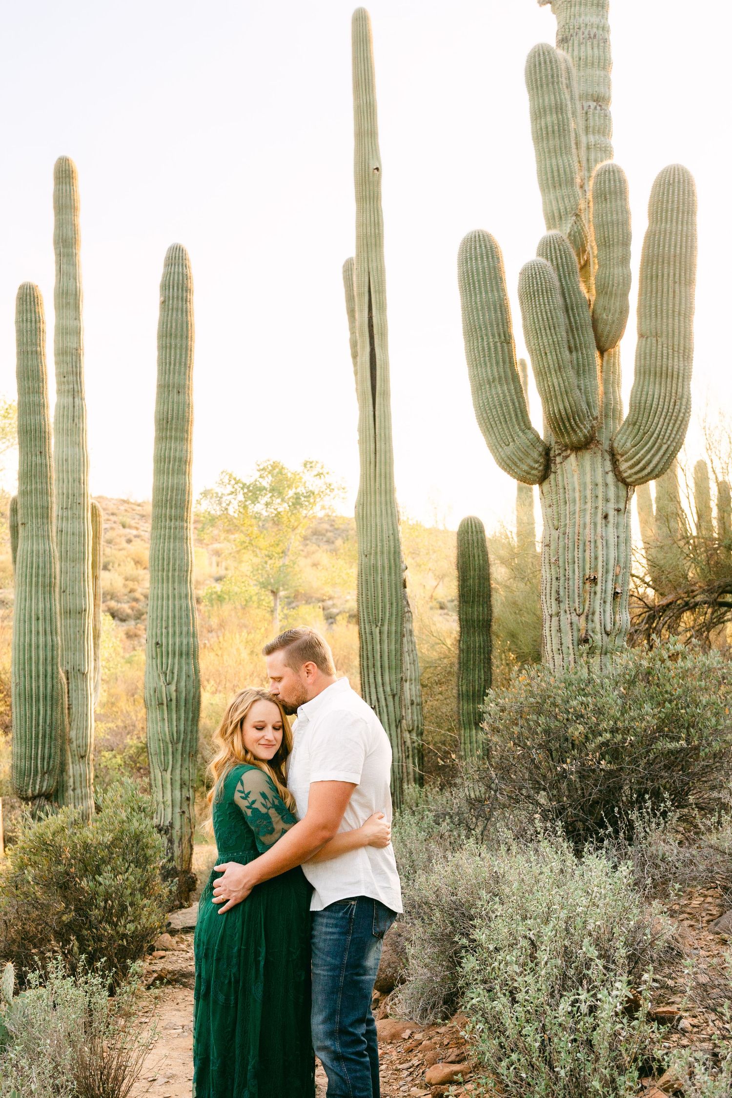 A pregnant woman smiling in a green dress has one hand on her belly and the other in her hair. She's surrounded by cactus and a rock path.