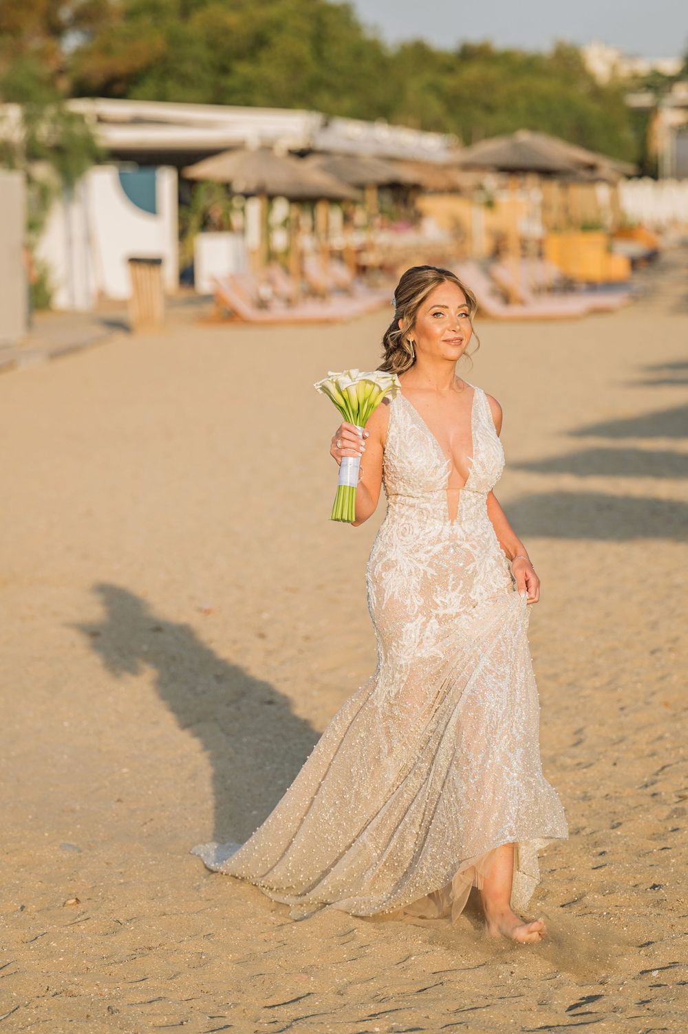 A bride in a lace wedding dress walks barefoot along a sandy beach at sunset holding a bouquet of flowers.