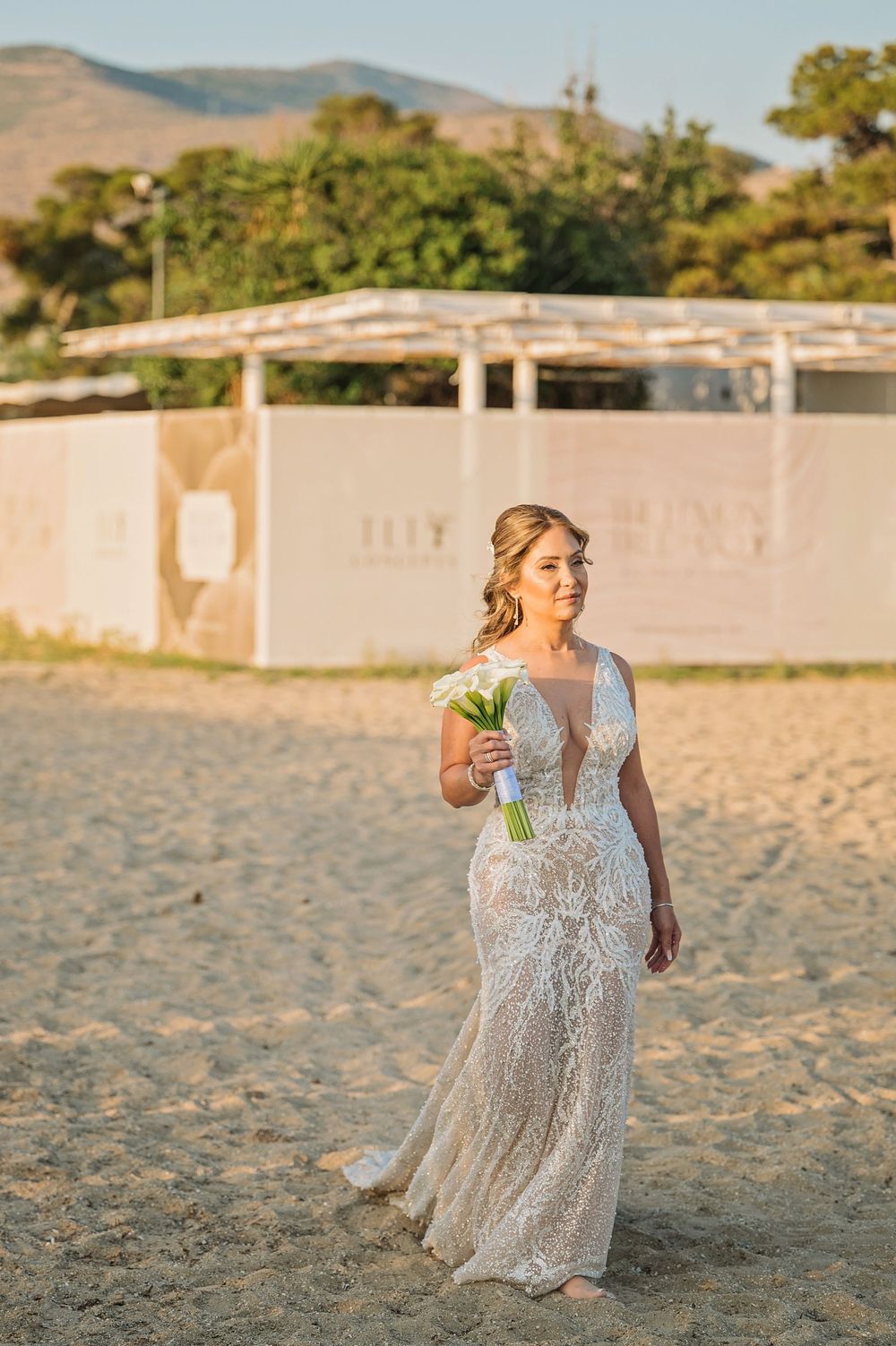 A bride in an elegant lace gown stands on a beach with mountains in the background holding white flowers.