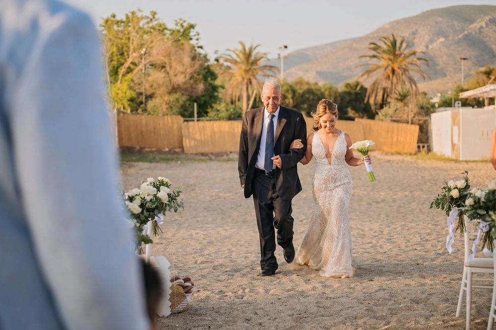A wedding processional on a sandy beach with mountains and palm trees in the background at sunset.