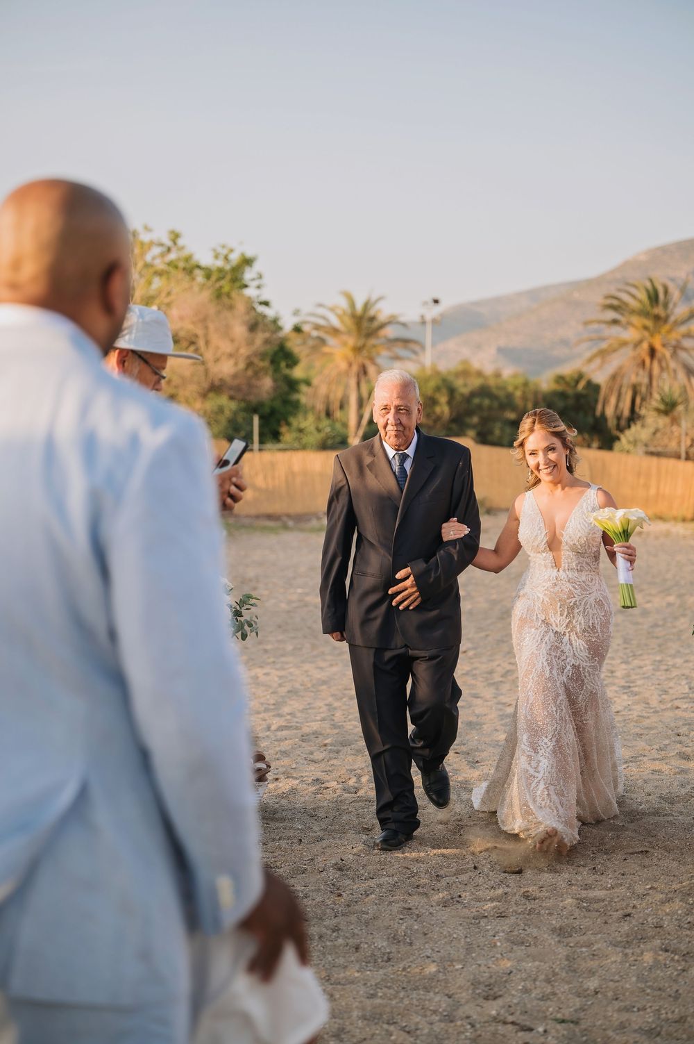 A wedding ceremony procession walking along a sandy beach path with palm trees in the distance.