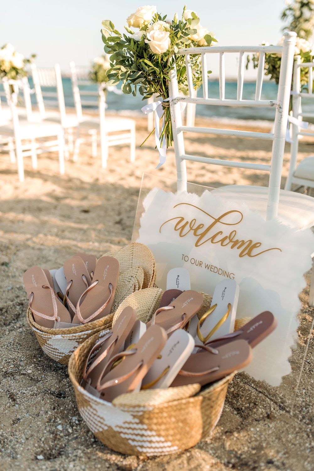Welcome sign and basket of flip-flops for beach wedding guests set on sandy shore.
