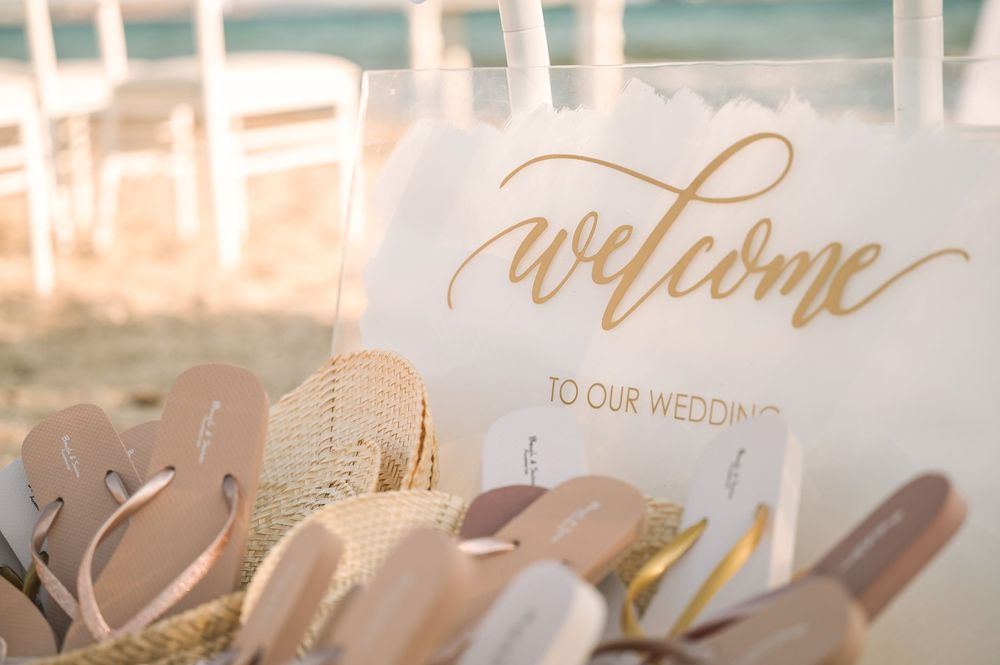 Close-up of golden 'Welcome to our wedding' sign with beach sandals in woven baskets.