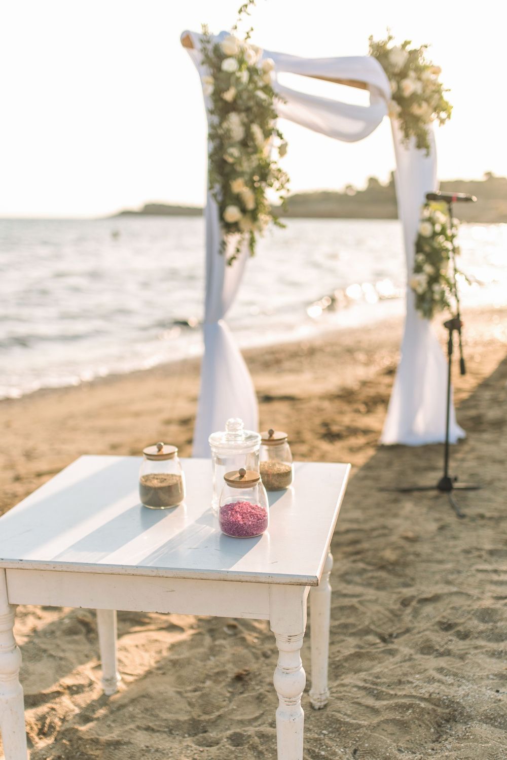 White wedding arch and decorative table with jars set up on beach at sunset.