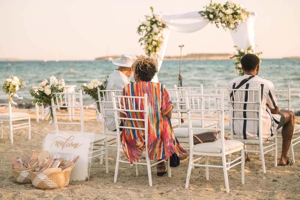 Casual beach wedding ceremony setup with guests seated in white chairs by the ocean.