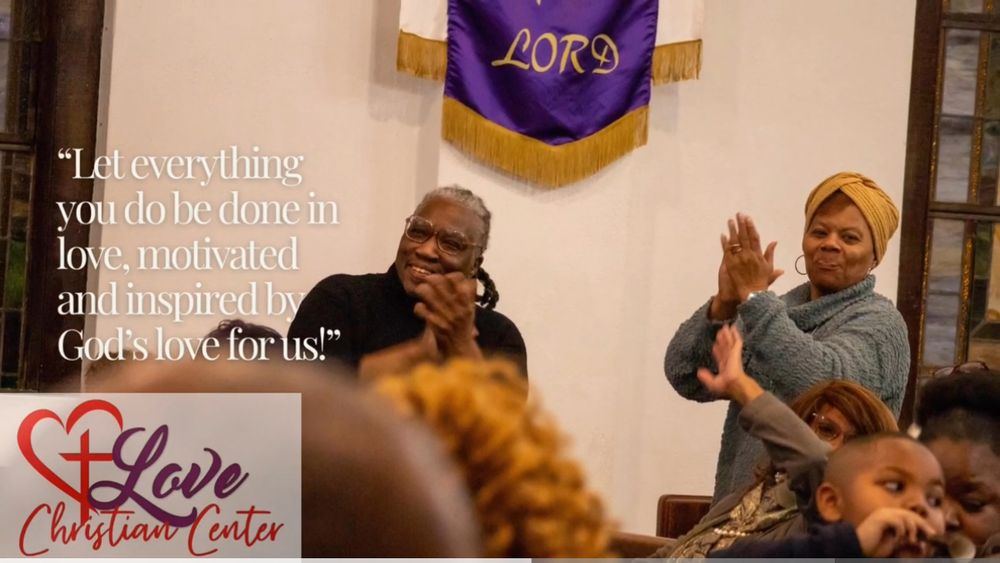 Two people stand worshipping in church under a purple and gold Lord banner during a Christian service.