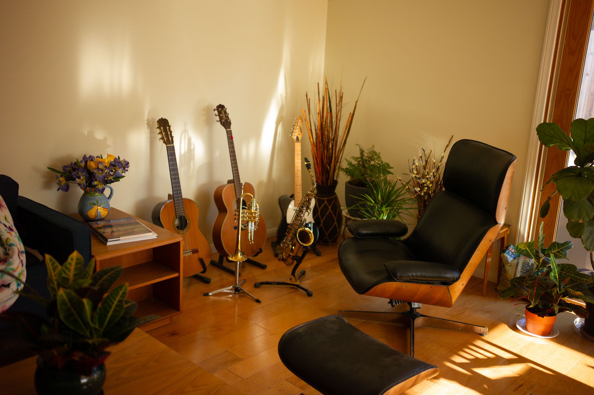 A cozy room corner with guitars on stands, house plants, and a comfortable black chair illuminated by warm lighting.
