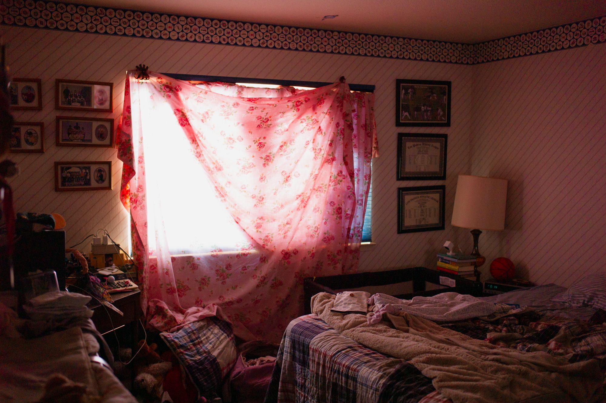 A messy bedroom with pink curtains filtering sunlight through the window and framed photos on the wall.