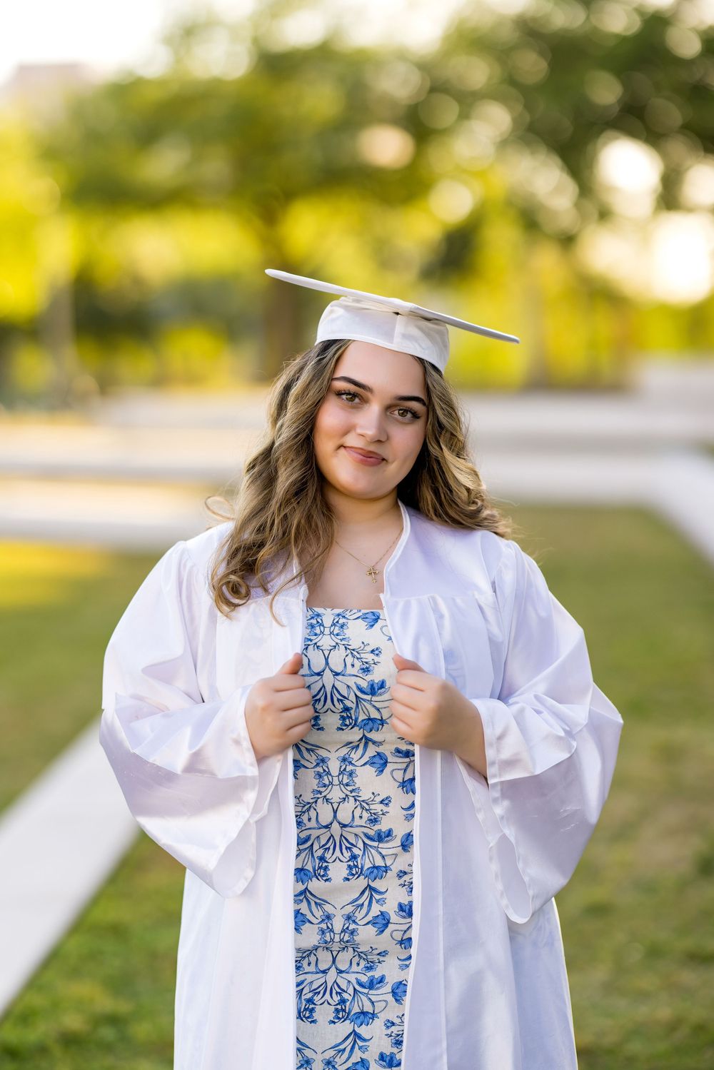 Graduate in white gown and blue floral dress poses outdoors during sunset.