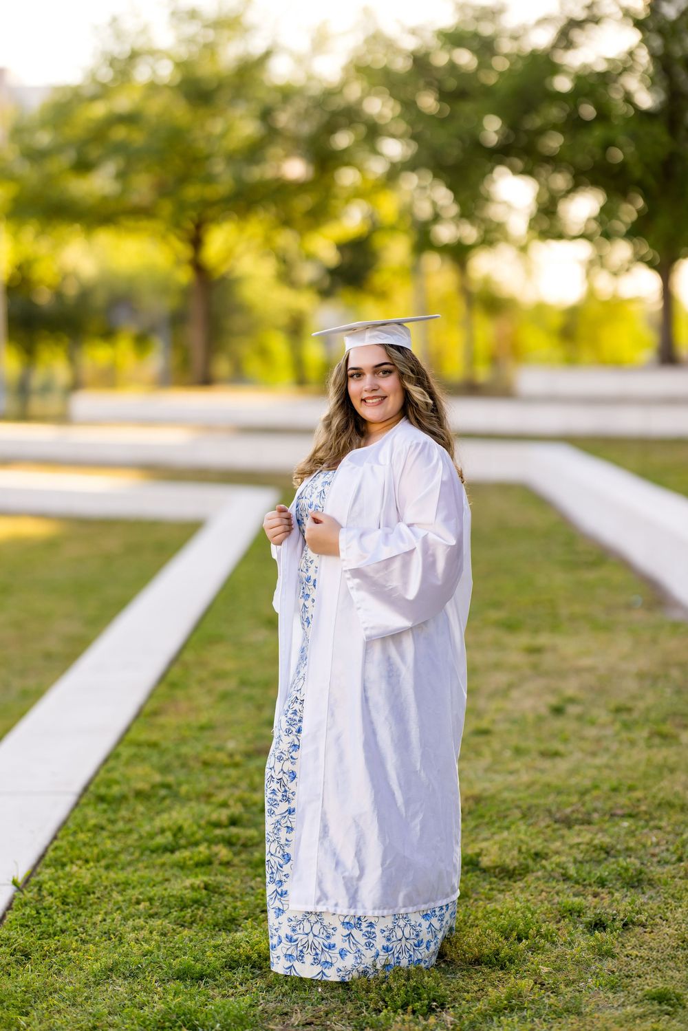 Graduate in white gown posing playfully on grass during sunset photoshoot.