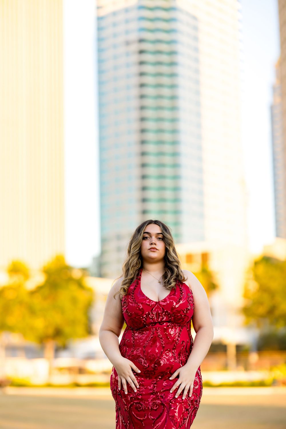 A red sequined evening gown is worn against an urban backdrop with a tall modern building and autumn trees.