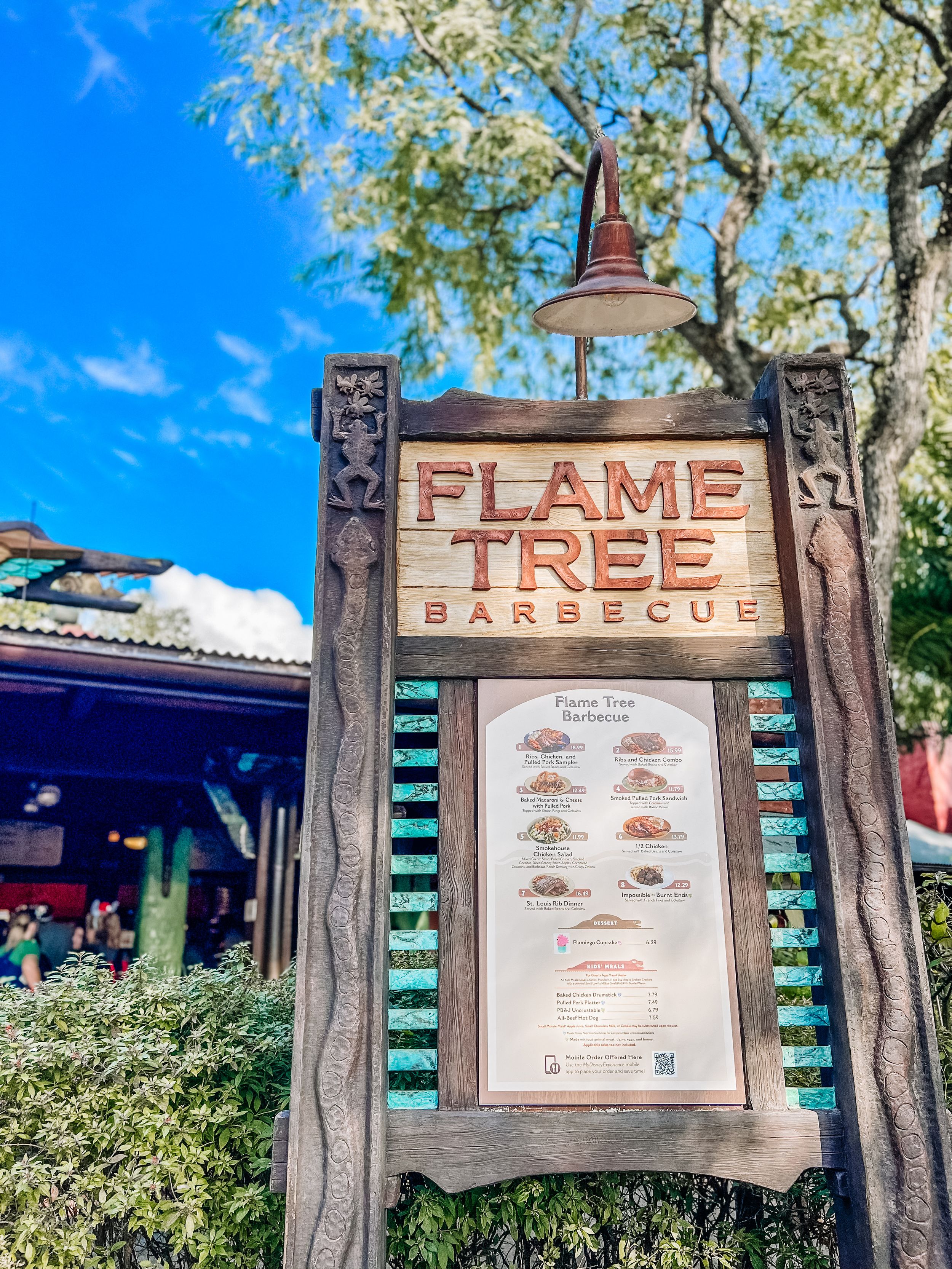 A rustic wooden menu sign for Flame Tree Barbecue restaurant with decorative carved posts against a bright blue sky.