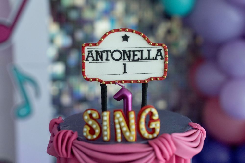 Close-up of a pink theater cake topper with SING and Antonella text in marquee style.