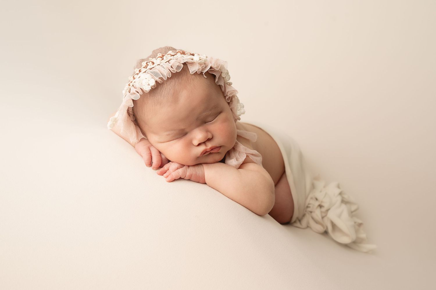 Sleeping newborn wearing pink lace bonnet posed on cream backdrop in artistic portrait.
