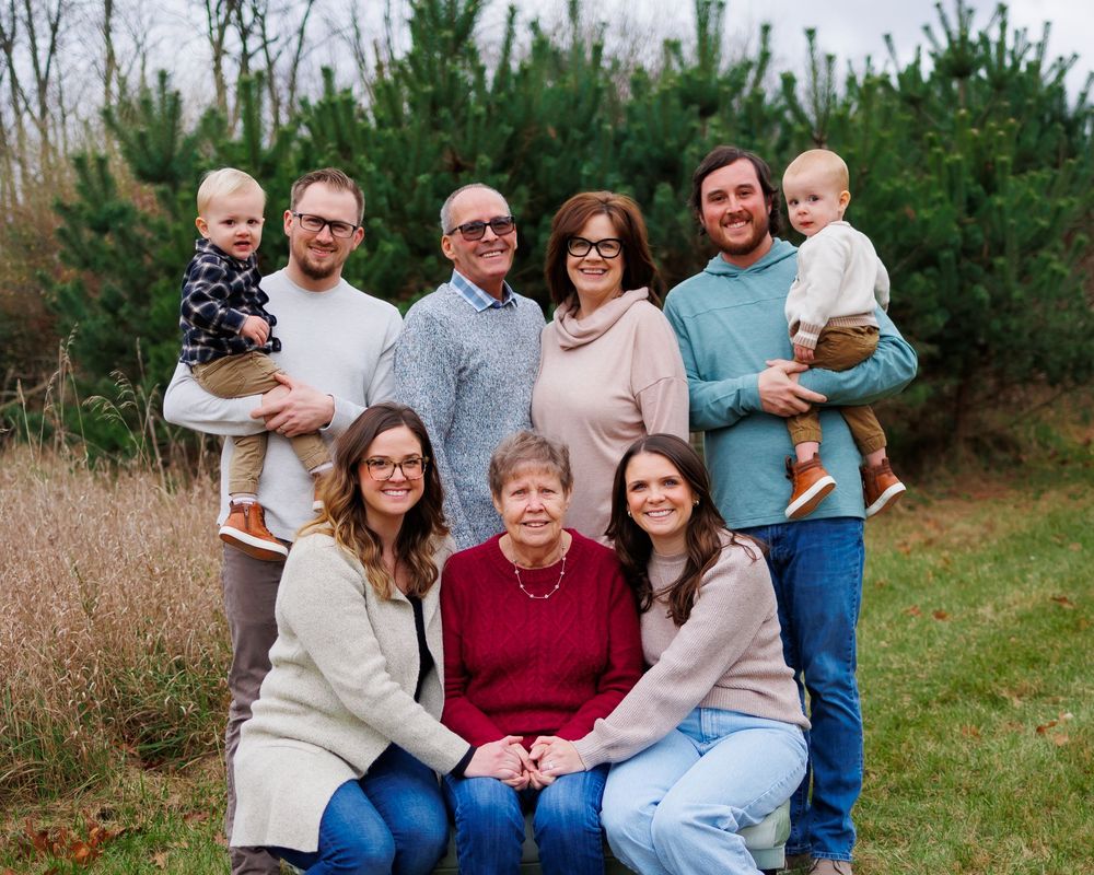 A multi-generational family poses together outdoors against a backdrop of evergreen trees and tall grass.