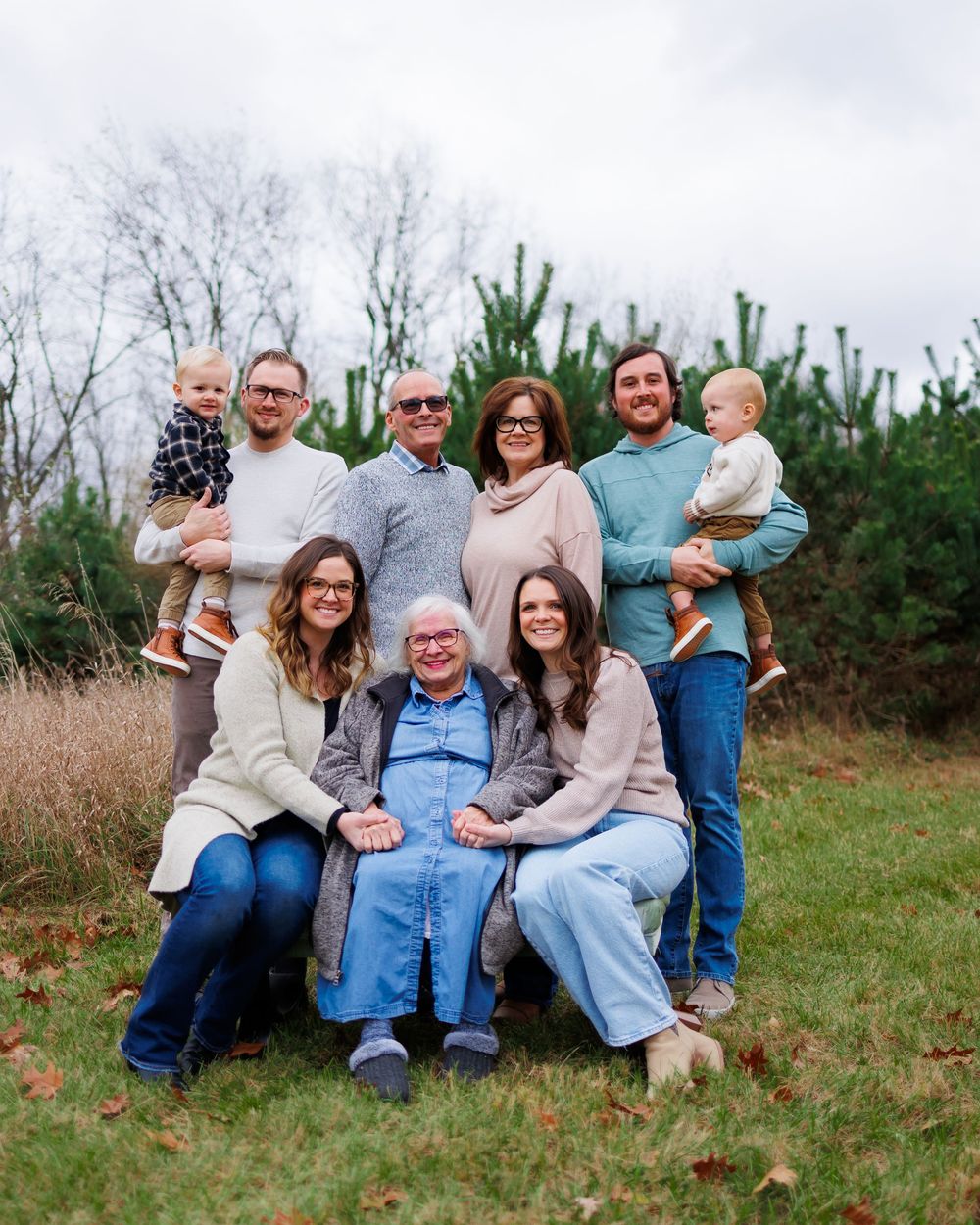 Multi-generational group sitting on a grassy hillside with evergreen trees in the background on an overcast day.