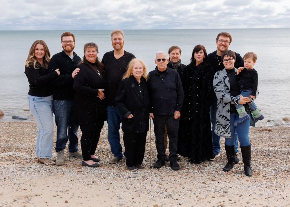 Group wearing black clothing poses together on a beach with the ocean and cloudy sky in the background.