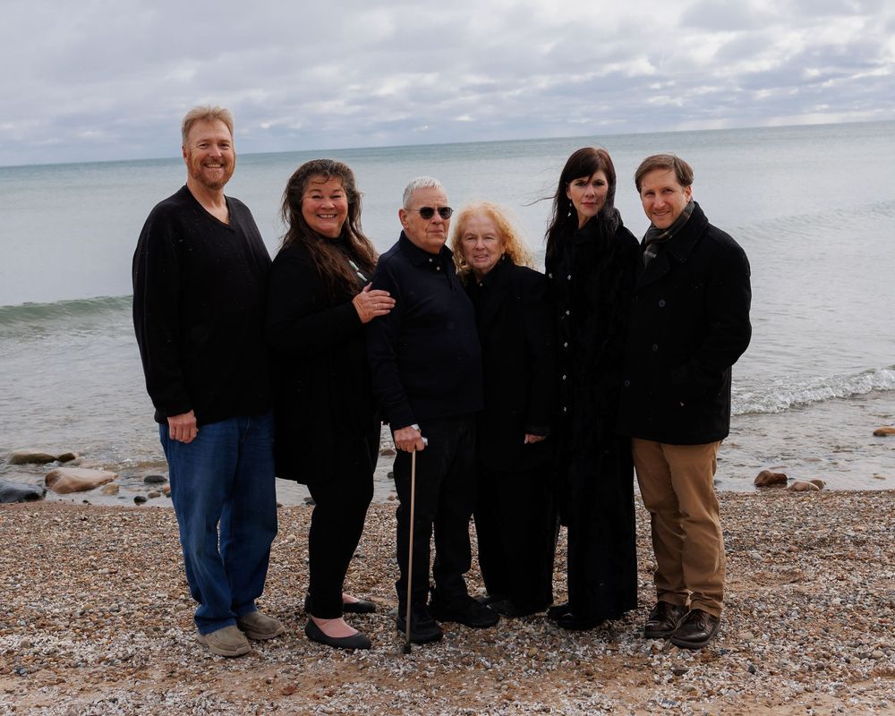 A group of six people wearing dark winter coats stand together on a rocky beach shore with cloudy skies and water behind them.