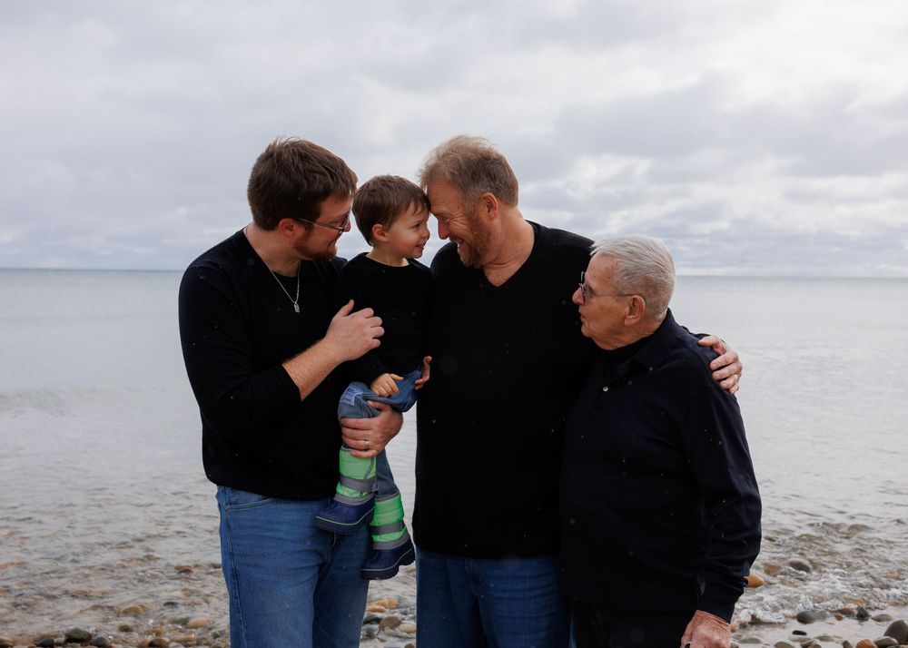Four people wearing dark shirts stand together and share a moment by the ocean shore on a cloudy day.
