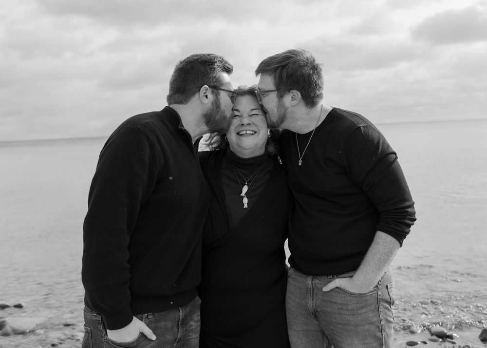 Black and white photo of three people in dark sweaters embracing and sharing a joyful moment on a beach.