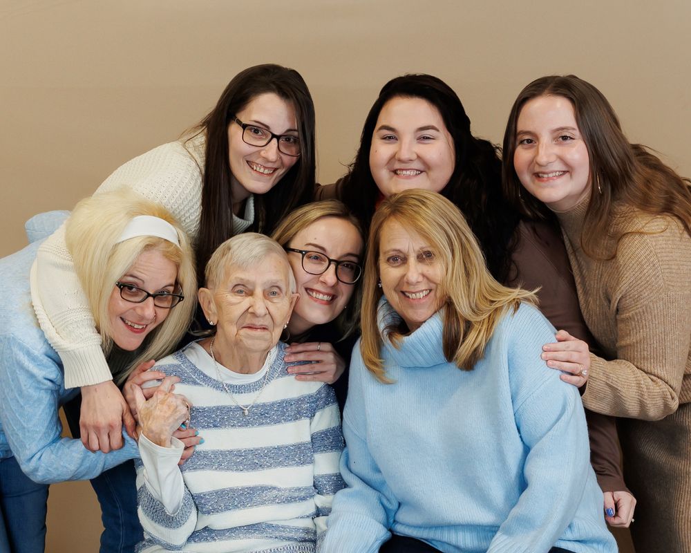 A multi-generational family group wearing casual attire smiles together for a portrait against a neutral background.
