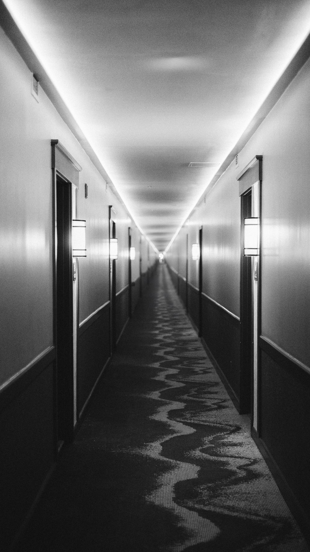 Long empty hotel corridor with stark white lighting and patterned carpet creating an eerie atmosphere.