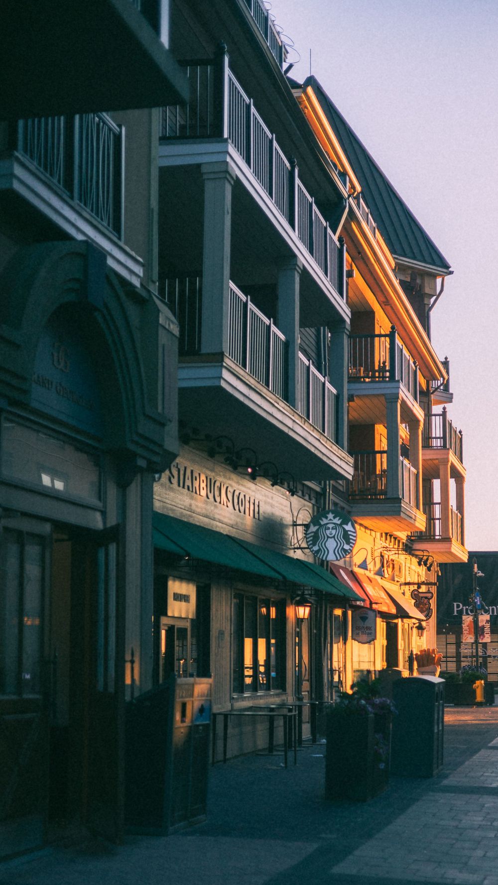 Storefronts along a quiet shopping street at twilight with warm glowing lights.