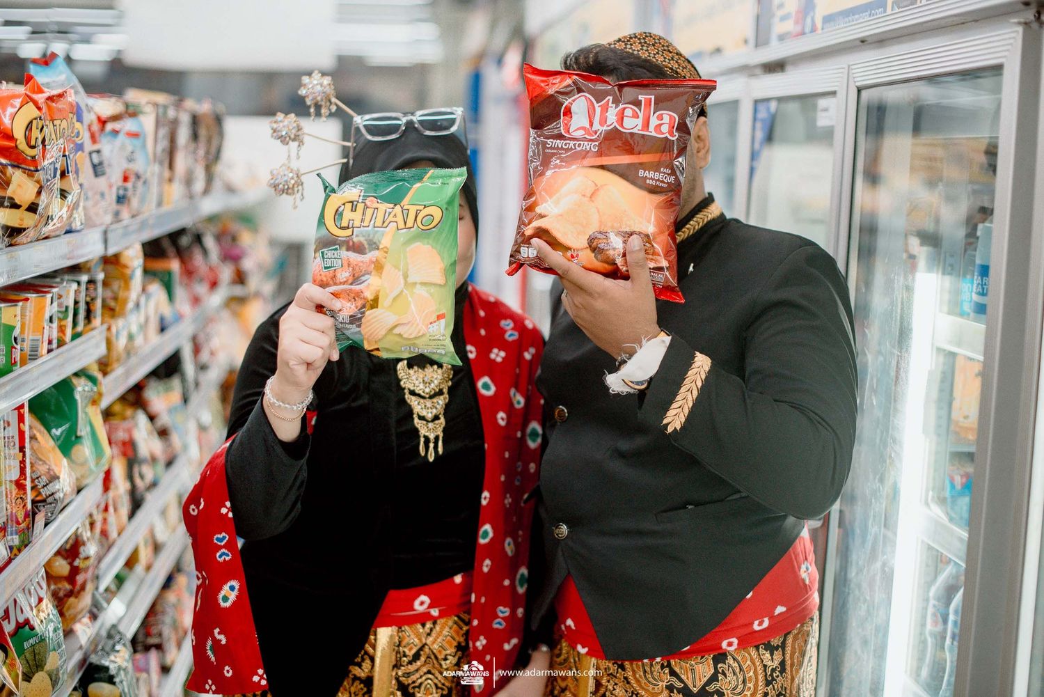 Two people in bandanas hiding behind snack bags in a grocery store aisle next to refrigerated cases.