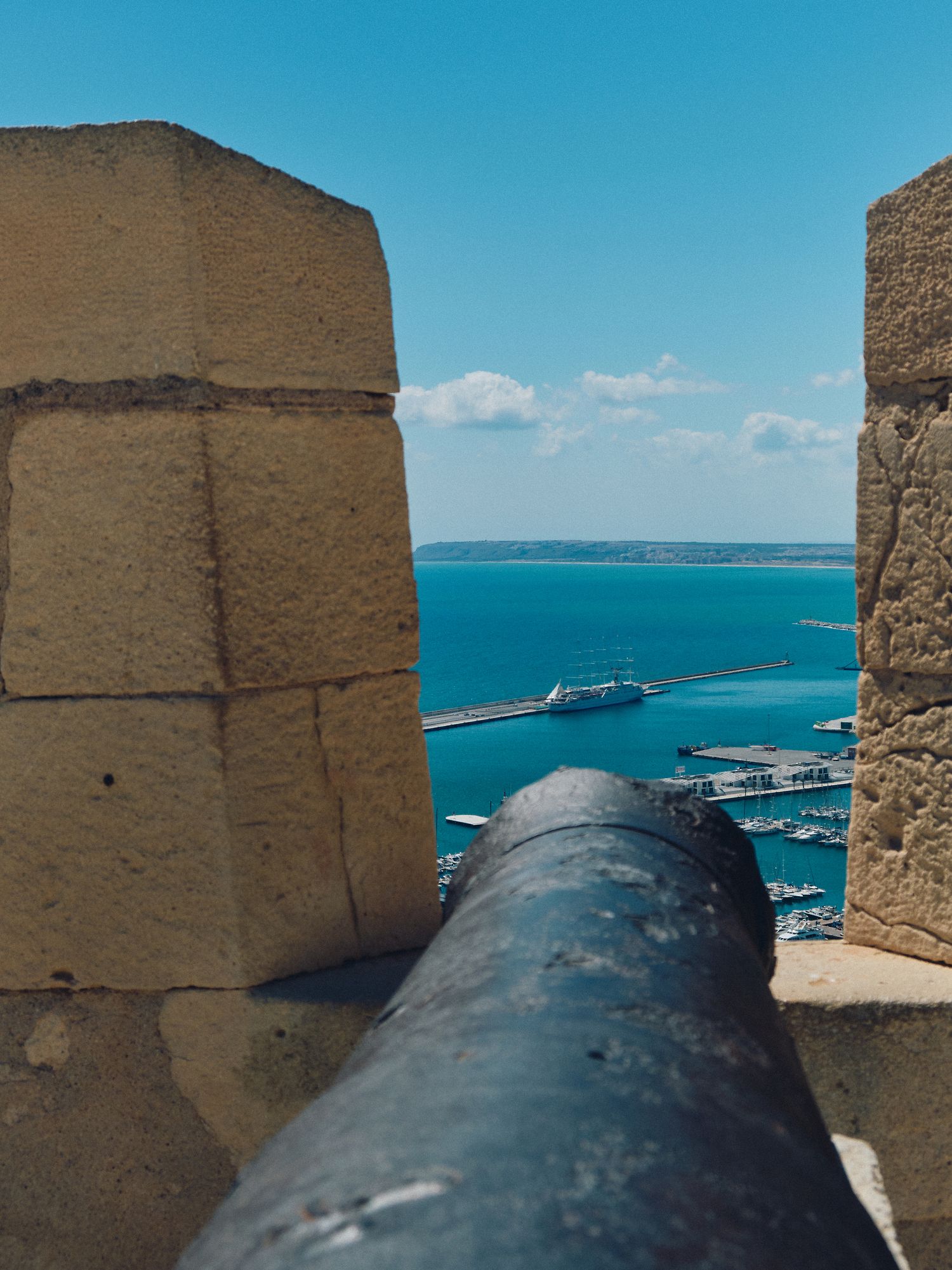 Ancient stone fortification with cannon aimed at turquoise harbor waters and distant bridge in Mediterranean setting.