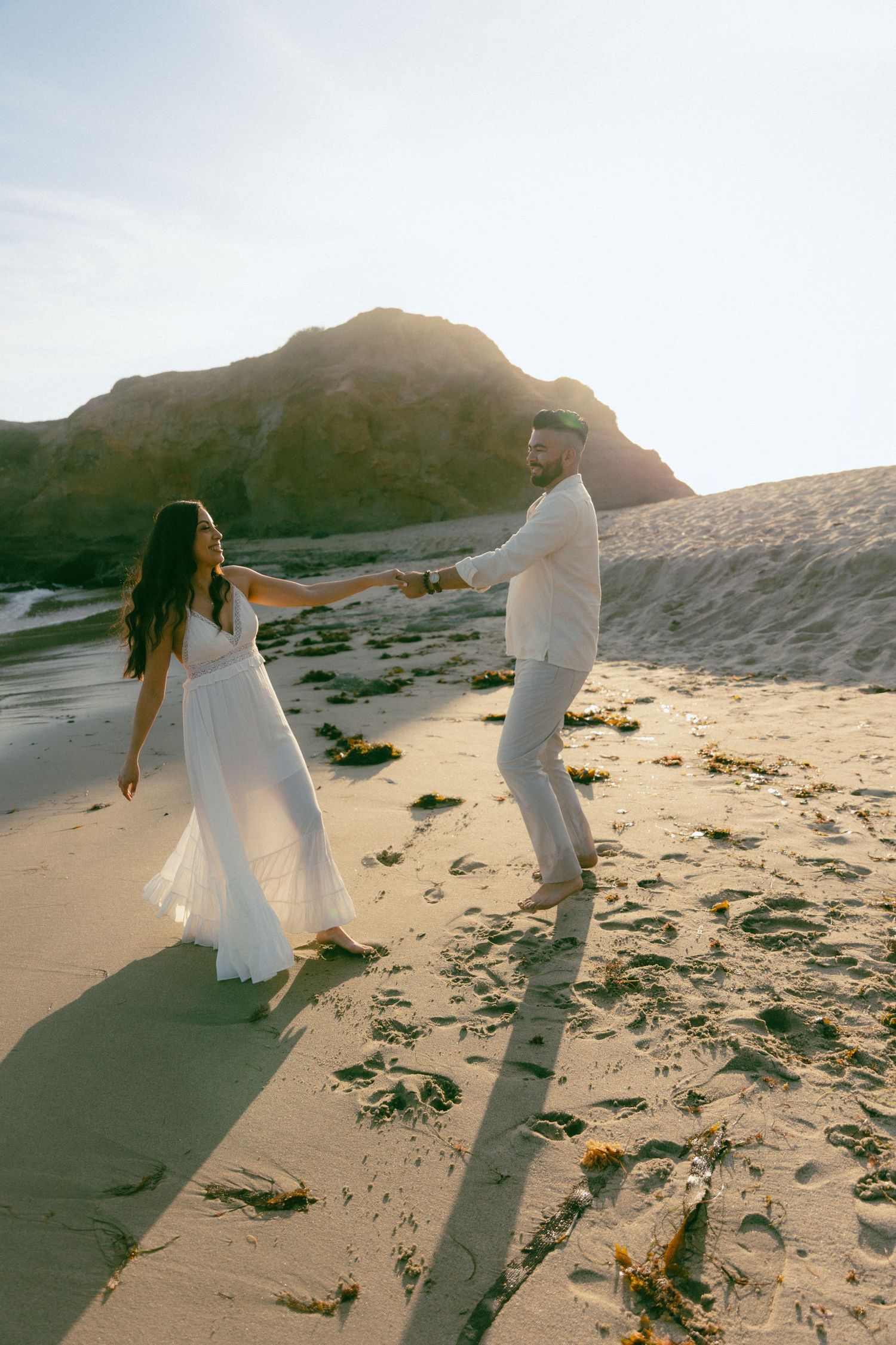 Two people in white outfits hold hands and dance together on a beach at sunset with mountains in the background.