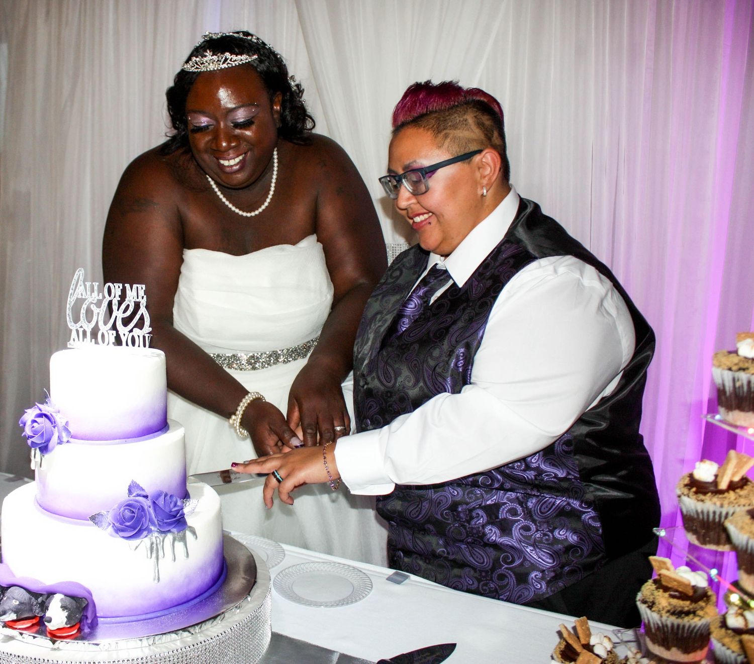 Happy newlyweds cutting their purple and white wedding cake together at reception.