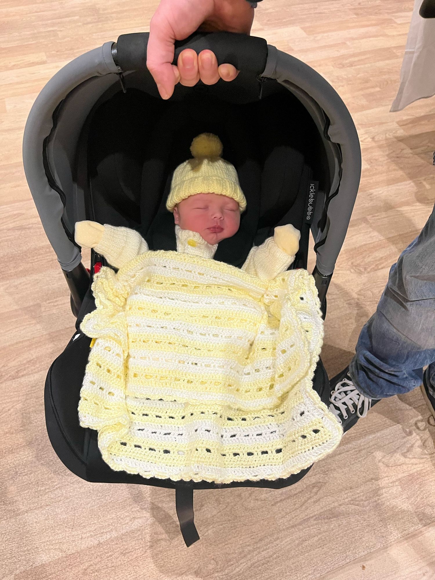 A baby wearing a yellow knitted dress and matching yellow hat sleeps peacefully in a black car seat.