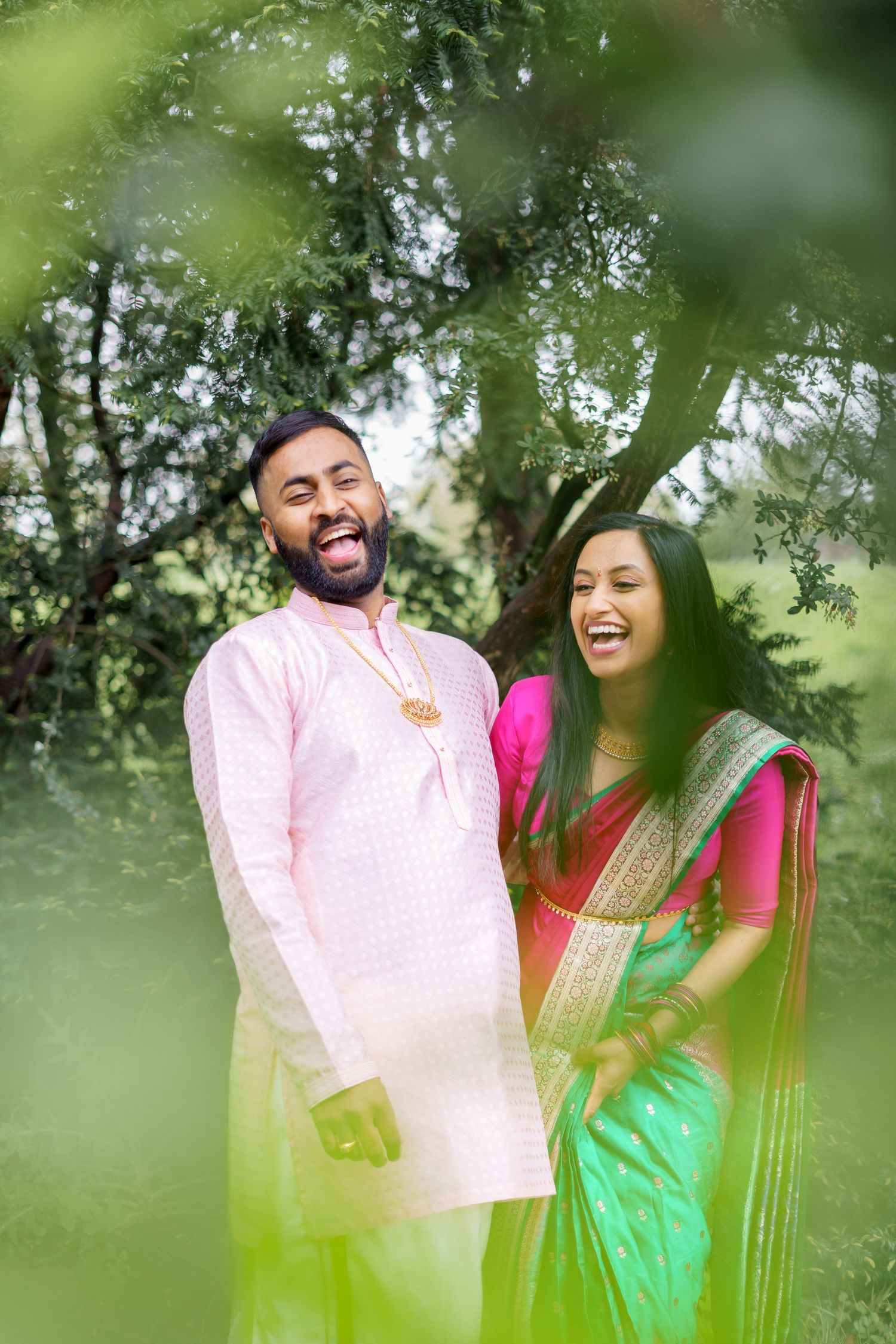 A couple in traditional Indian attire share a joyful moment together outdoors, framed by green foliage.