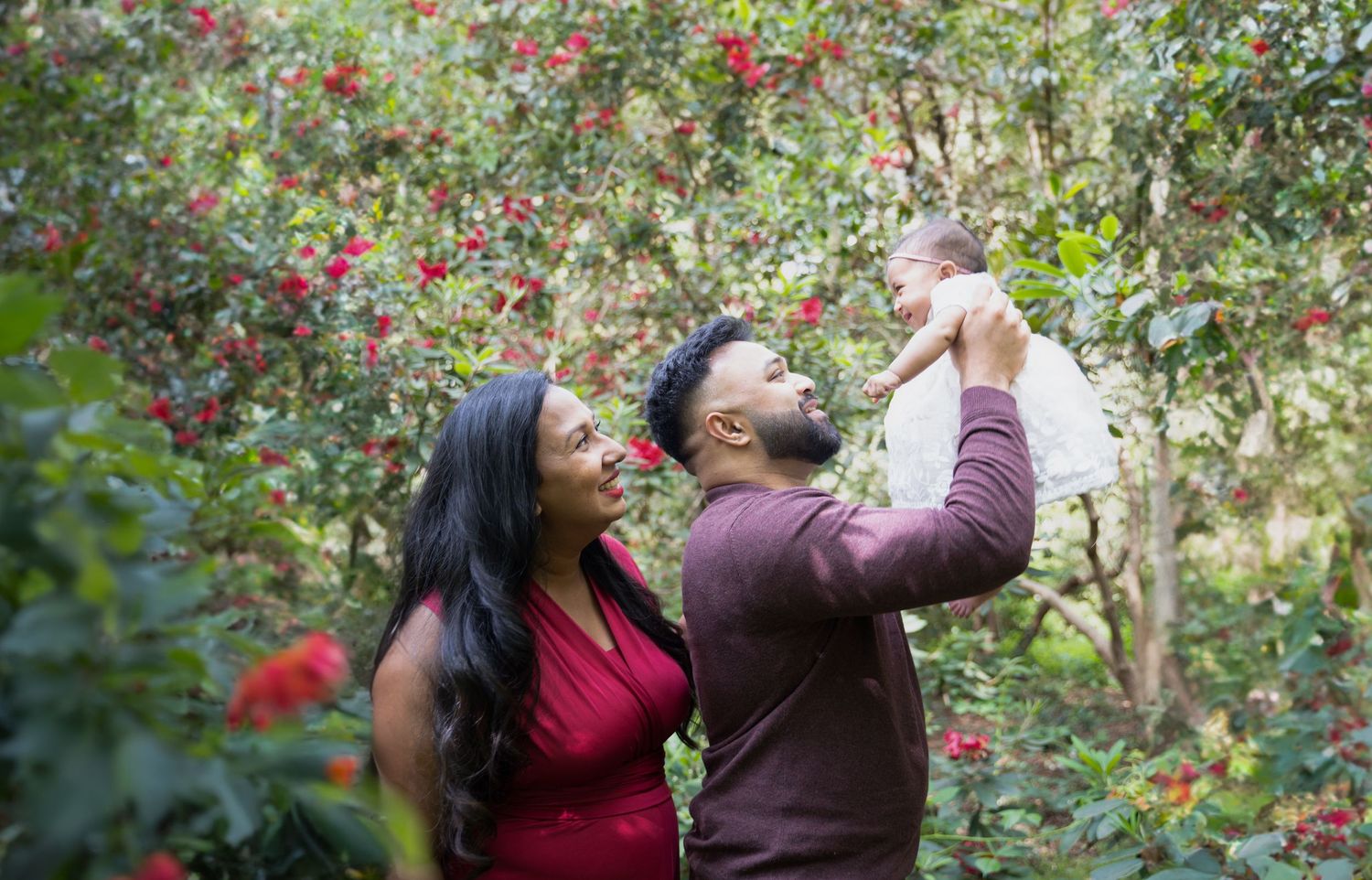 A couple enjoys time together in a garden with their baby beside blooming red flowers and lush greenery in the background.