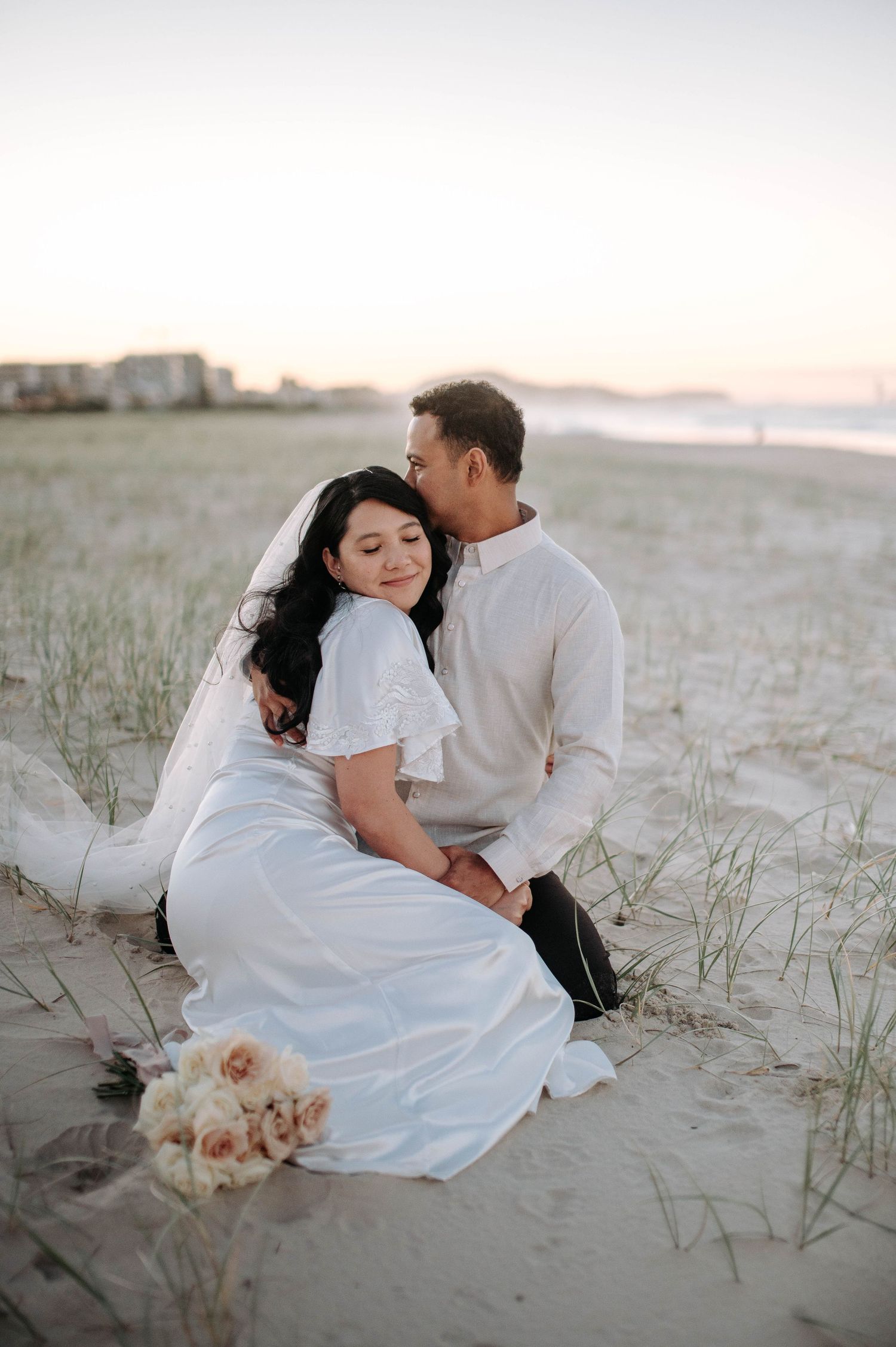 Romantic wedding couple sits together on sandy beach dunes at sunset with bouquet nearby and soft lighting.