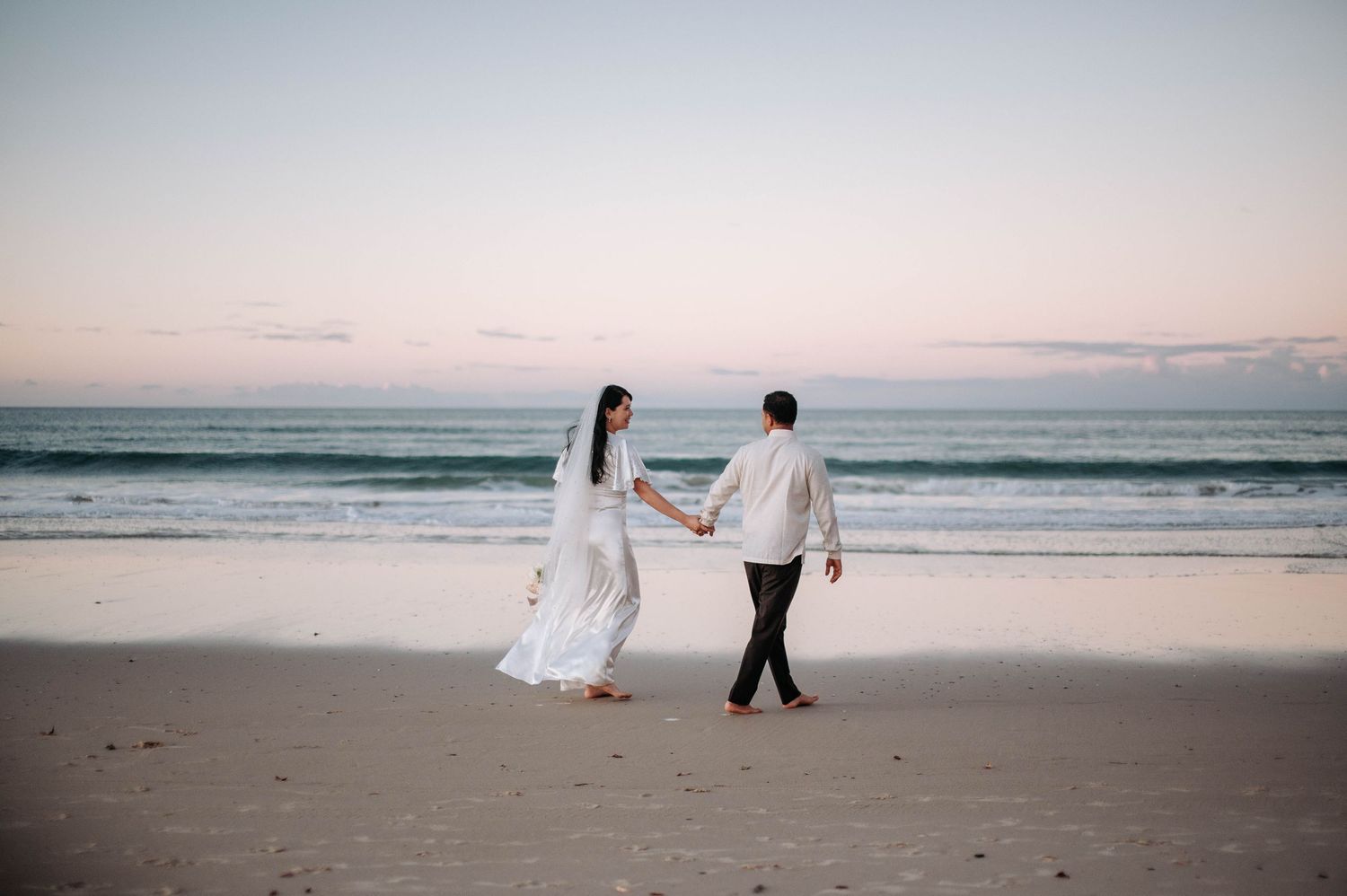 A couple in wedding attire walks hand in hand along a beach at sunset with ocean waves in the background.