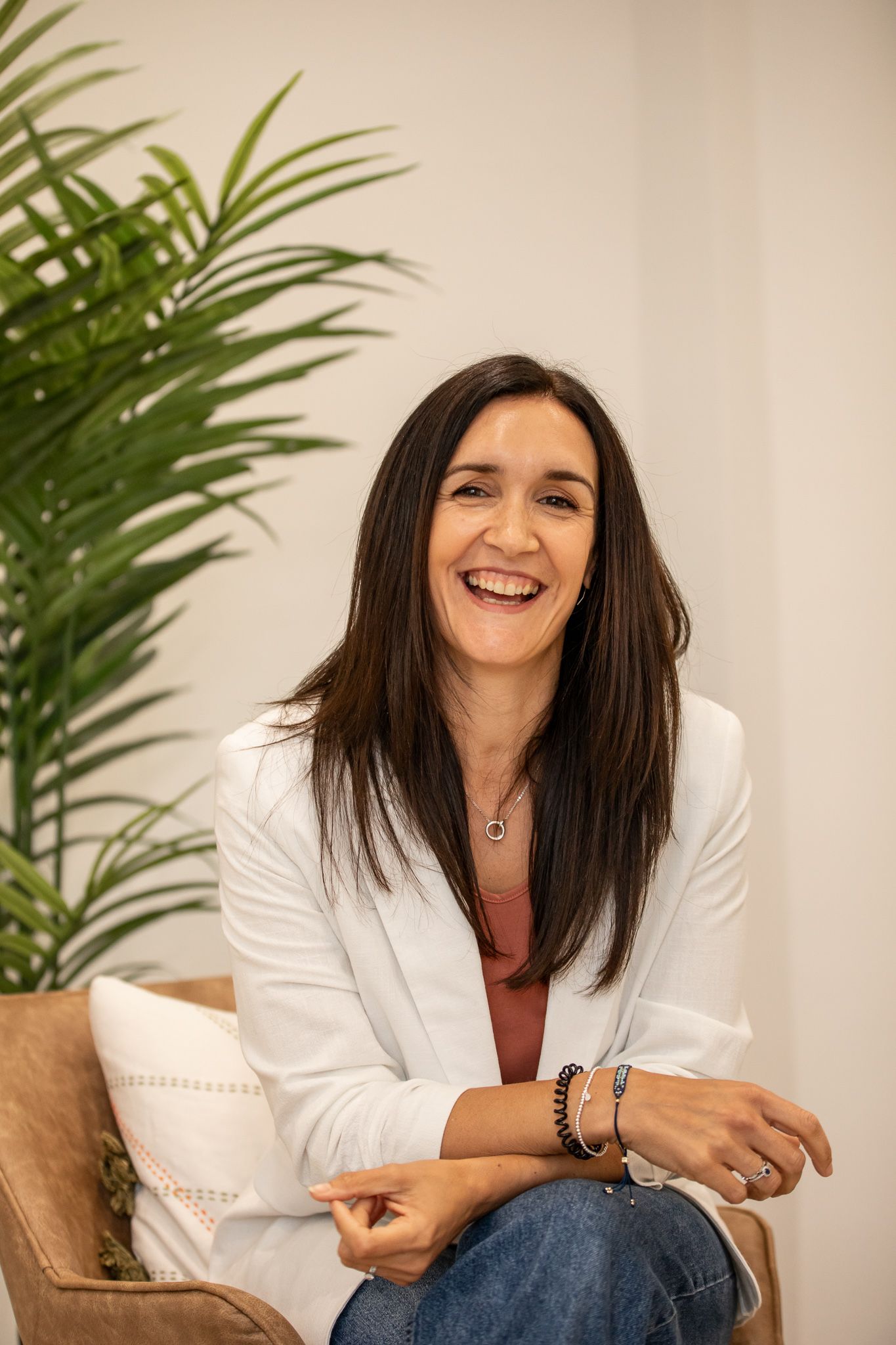 Professional headshot of someone in a white blazer and rust-colored top sitting casually by a tropical plant.