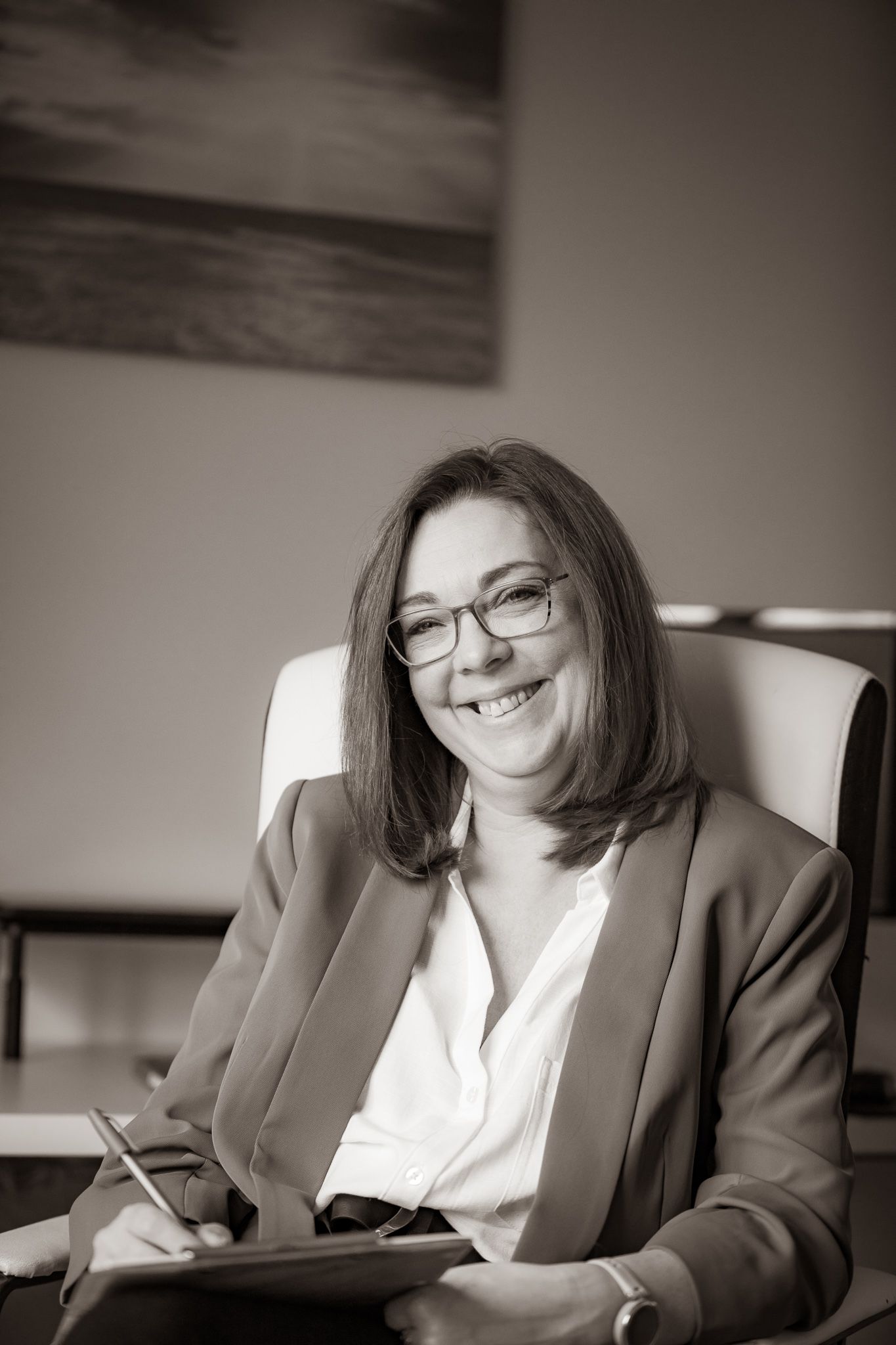 Professional wearing glasses and a blazer sits in an office chair with notepad, smiling warmly in black and white portrait.