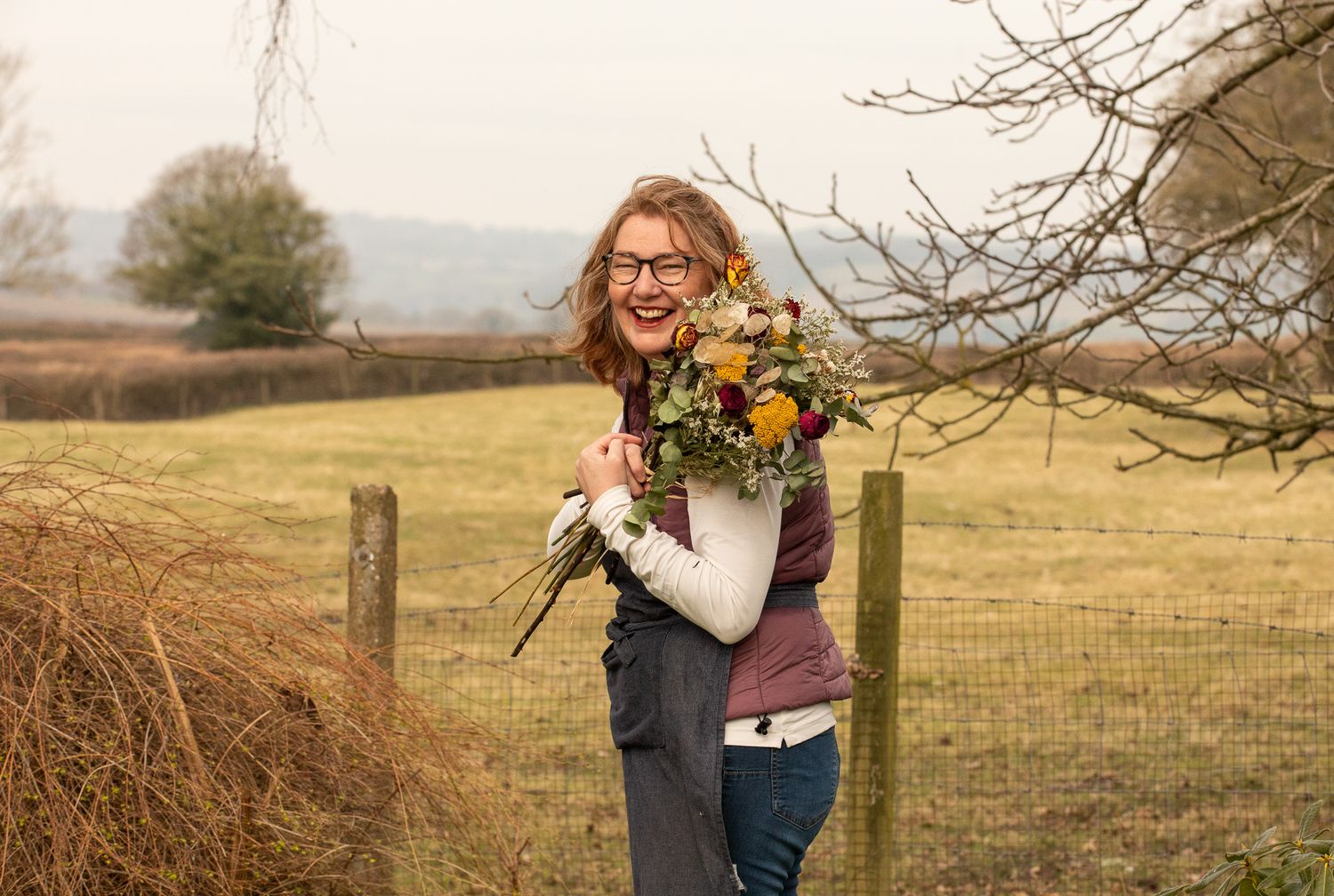 Someone holds a vibrant bouquet of yellow and orange flowers while standing by a rustic wooden fence in a rural field.