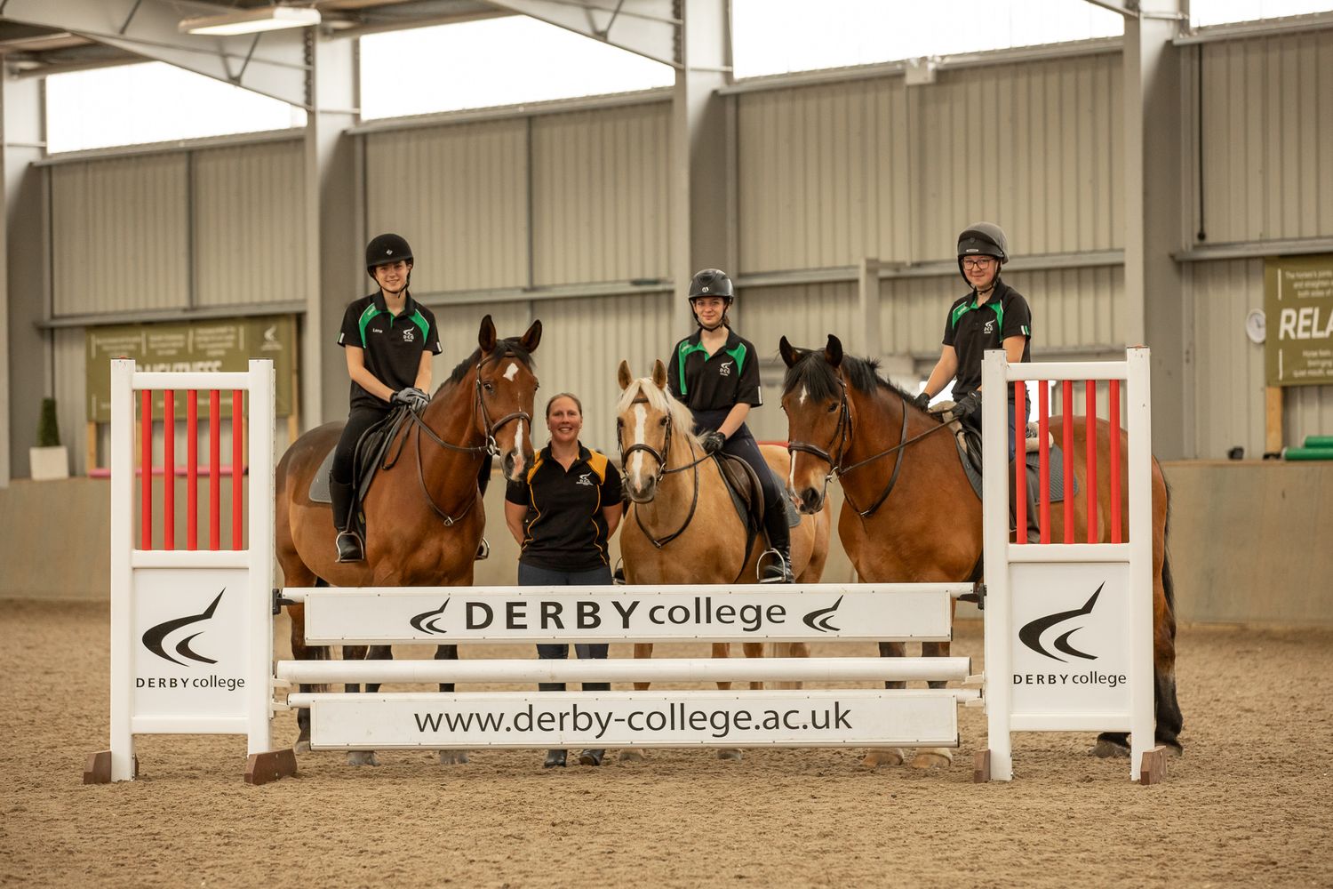 Three equestrians on horseback jumping over a Derby College branded obstacle in an indoor riding arena.