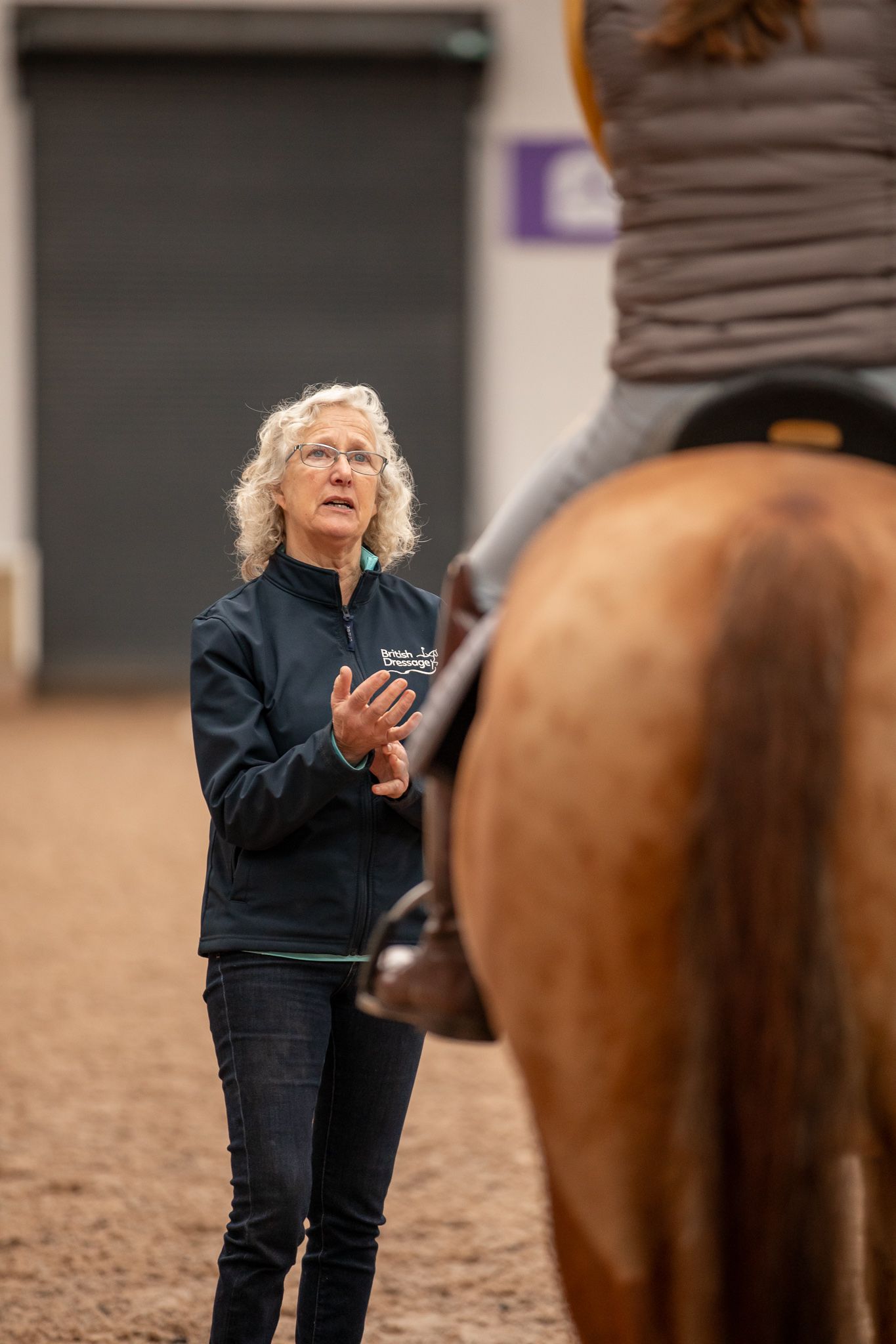 Riding instructor in navy jacket gives instruction to mounted student in indoor arena during equestrian lesson.