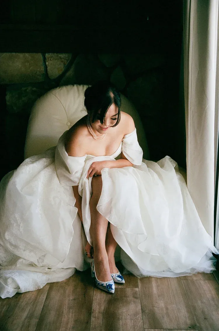 A bride in a white wedding dress adjusts her teal blue shoes while sitting on a wooden floor.