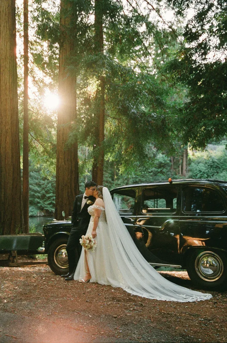 A classic black vintage car parked in a redwood forest creates a romantic wedding photo backdrop at sunset.