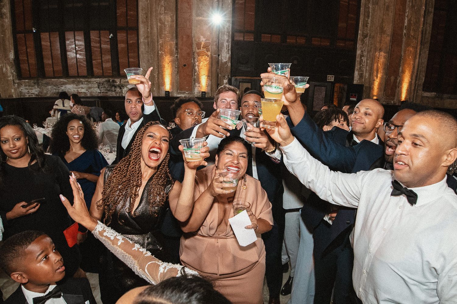 Wedding guests dance and celebrate with raised drinks at a lively reception party.