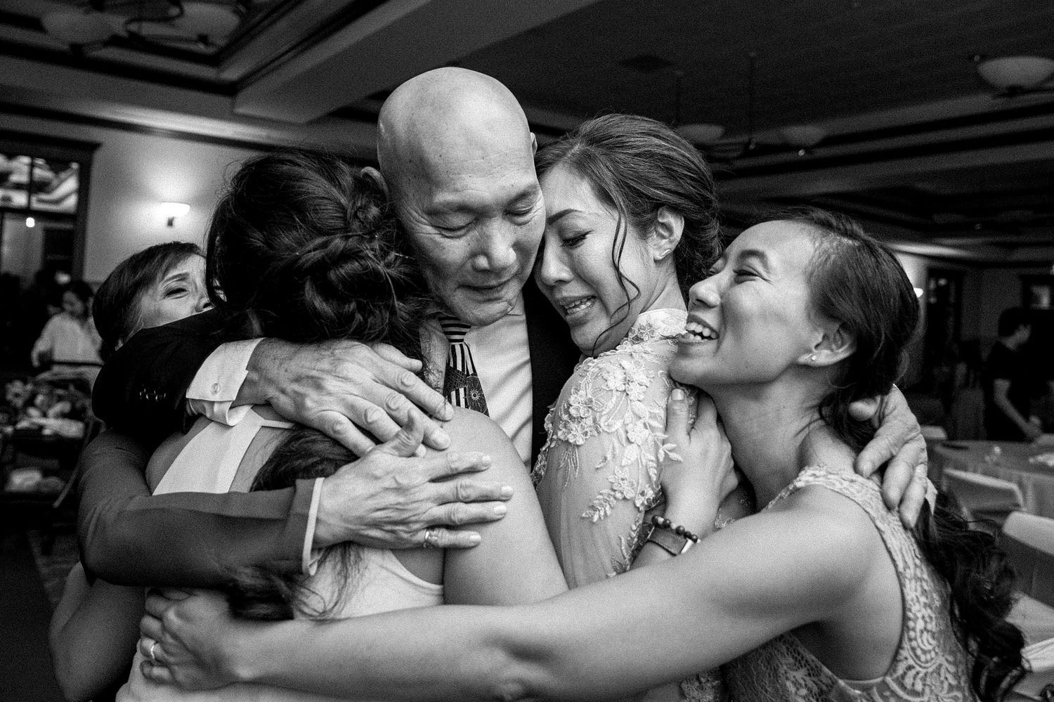 A candid black and white photograph captures an emotional group hug at a wedding reception.