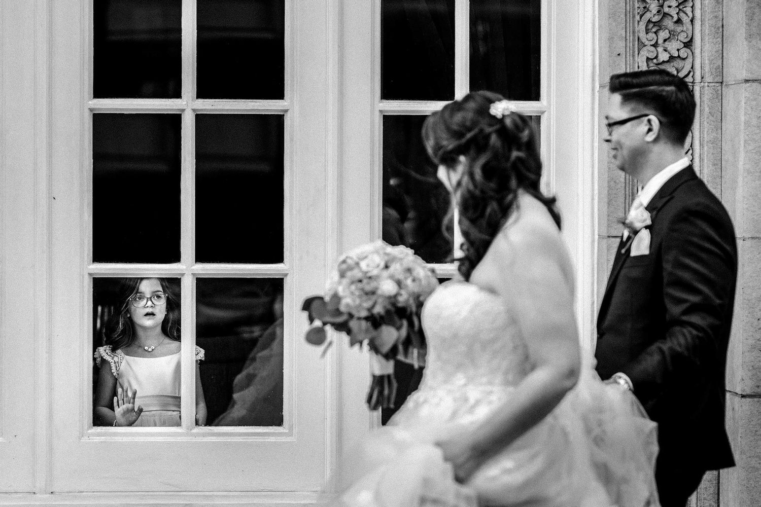 Black and white wedding photograph showing a bride and groom outside with a reflection visible in a window.