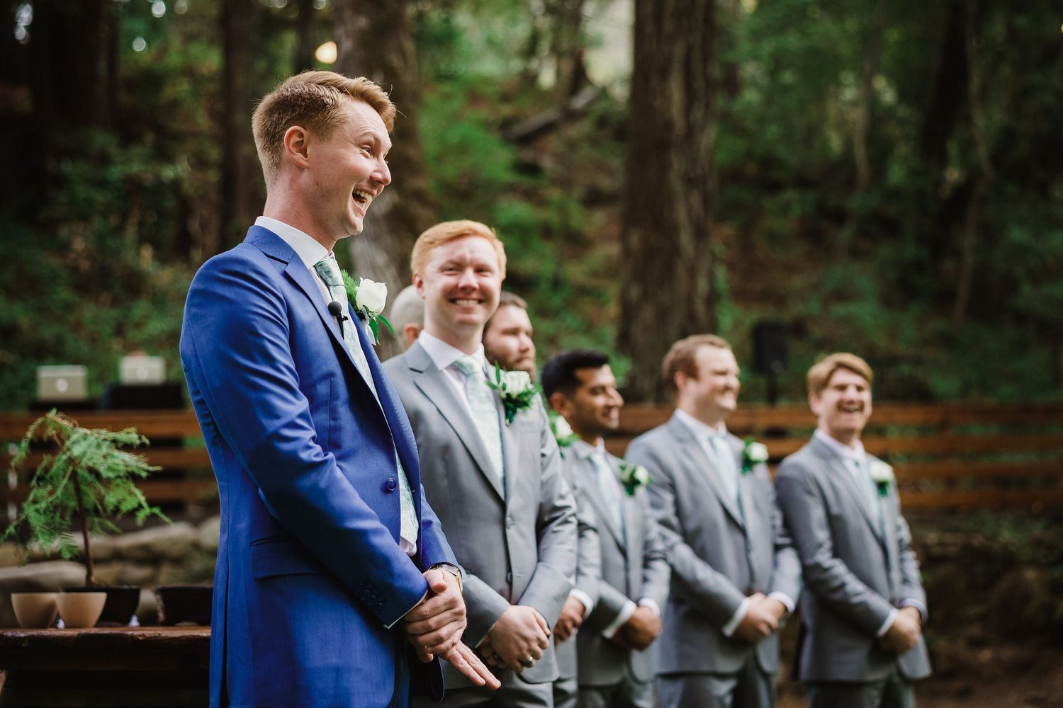Wedding party members in gray and blue suits share a candid laughing moment during an outdoor celebration.